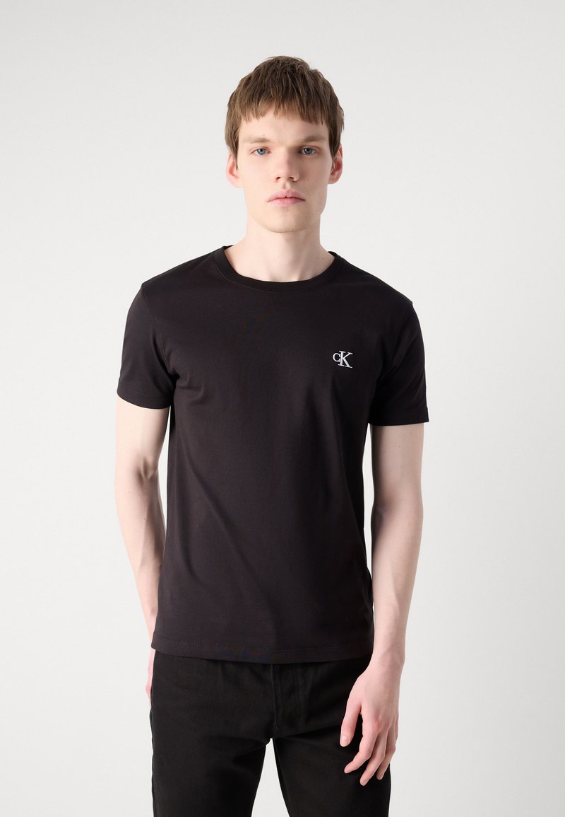 Joven de pie de frente vistiendo una camiseta negra Calvin Klein y pantalones negros contra un fondo claro y liso.