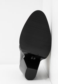 Zapato de tacón alto negro con puntera afilada y suela texturizada. Presenta un tacón curvado y el nombre de la marca en relieve a lo largo del borde.