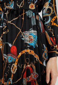 Vestido de tela negra con patrones coloridos de cadenas, cuerdas y herrajes decorativos, con textura plisada y mangas anchas.