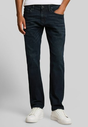 Mann trägt dunkelblaue Straight-Leg-Jeans und weiße Sneakers, mit einer Hand in der Tasche vor grauem Hintergrund.