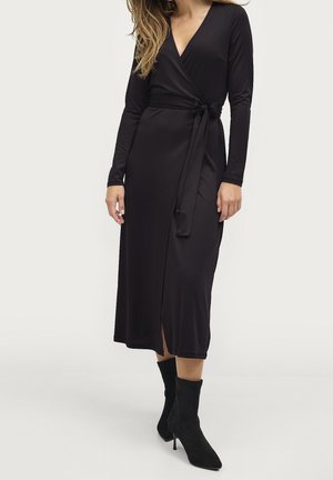 Kobieta w czarnej, długorękawowej sukience kopertowej z paskiem związanym w talii i czarnych botkach na wysokim obcasie, stojąca na jednolitym tle.
