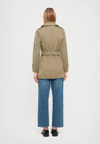 Lichtgewicht, gestreepte jas in olijfgroen met een ceintuur in de taille en een klassieke kraag, gedragen met blauwe wijde jeans en zwarte schoenen.