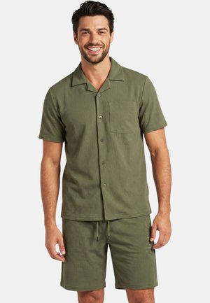 Homme souriant portant une chemise boutonnée manches courtes couleur olive avec poche poitrine et un short assorti à cordon, sur fond blanc.