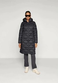 Γυναίκα που φοράει ένα μακρύ μαύρο παραγεμισμένο παλτό με κουκούλα, σκούρα παντελόνια, άσπρα αθλητικά παπούτσια και μεγάλα σκούρα γυαλιά ηλίου, στέκοντας μπροστά σε απλό φόντο.