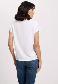 Biała koszula z krótkim rękawem o okrągłym dekolcie, gładka w dotyku, w luźnym kroju, w połączeniu z niebieskimi dżinsami. Prosty i uniwersalny fason.