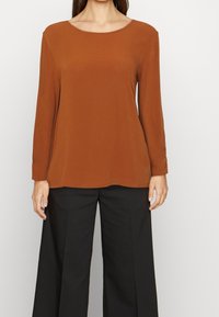 Maglietta a maniche lunghe marrone realizzata in materiale morbido, con scollatura ampia e vestibilità larga, abbinata a pantaloni neri a gamba larga.