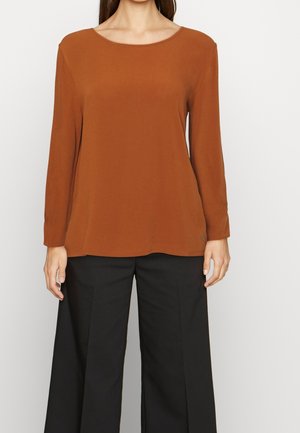 Maglietta a maniche lunghe marrone realizzata in materiale morbido, con scollatura ampia e vestibilità larga, abbinata a pantaloni neri a gamba larga.