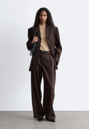 Kobieta z długimi kręconymi włosami, ubrana w oversize ciemnobrązowy garnitur, beżowy top, czarny pasek, czarne spiczaste buty i trzymająca czarną torebkę na ramię.