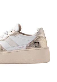 Hvide sneakers med metallisk guldaccent, tekstureret overdel, flad platformssål, sort logo-detalje og perforerede designelementer.
