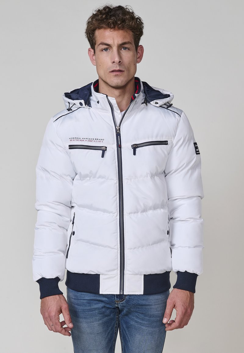 Chaqueta acolchada blanca con capucha, que cuenta con bolsillos con cremallera en el pecho, acentos en azul y puños acanalados. Textura suave, diseño acolchado.