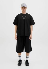 Zwarte oversized t-shirt met korte mouwen, zwarte shorts, grijze pet, zilveren ketting en zwarte sneakers met witte gestreepte sokken.