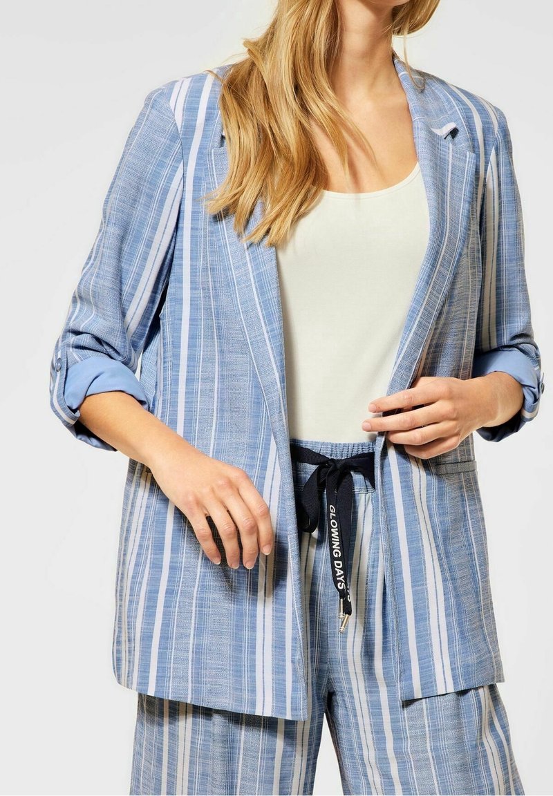 Femme portant un blazer à rayures verticales bleues et blanches avec un pantalon assorti et un haut blanc, tenant le revers du blazer et le cordon de la taille.