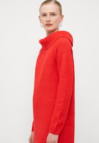 Abito rosso lavorato a maglia con scollo a ciambella, maniche lunghe e vestibilità comoda. Il materiale è morbido e presenta una sottile texture.