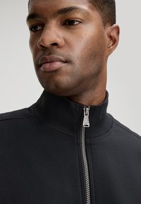 Zwarte jas met rits en een hoge kraag, gemaakt van textuurrijk stof. Voorzien van een metalen rits en contrasterende stikseldetails.