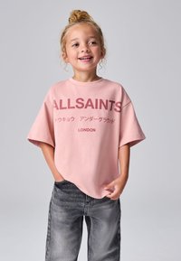 Vaaleanpunainen ylipitkä t-paita, jossa on teksti "ALLSAINTS", lyhyet hihat ja pyöreä pääntie, yhdistetty tummanharmaisiin farkkuihin. Yksinkertainen, rento muotoilu.