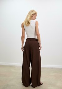 Femme blonde portant un haut sans manches blanc et un pantalon marron à jambes larges, se tenant debout et faisant face à l'extérieur dans un environnement intérieur minimaliste.
