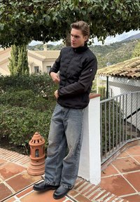 Jonge man in een zwarte jas en brede jeans die zijn mouw rechtzet, staand op een betegeld terras met groenblijvende planten en heuvels op de achtergrond.