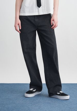 Persona vistiendo jeans negros de corte holgado, camisa blanca, corbata negra y zapatillas negras y blancas, de pie sobre un suelo alfombrado azul.