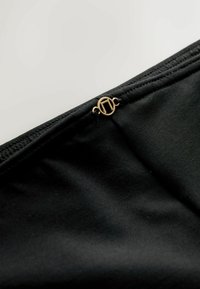 Mustast kangast, millel on sile tekstuur, ja peen kuldse tooniga logo kaunistus kinnitatud servasaumile.