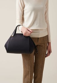Black structured handbag with textured fabric, two short handles, and gold hardware accents. Paired with patterned brown trousers and a light sweater.