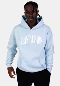 Hoodie azzurro chiaro con tasca frontale, maniche ampie e testo "CRYSTAL PARIS" in bianco. Tessuto morbido con una texture liscia.