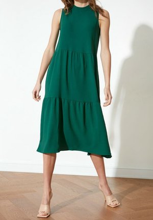 Ärmelloses dunkelgrünes Midikleid mit gestuftem Rock, getragen von einer stehenden Person mit hellbraunen offenen High-Heel-Sandalen auf einem Holzboden.