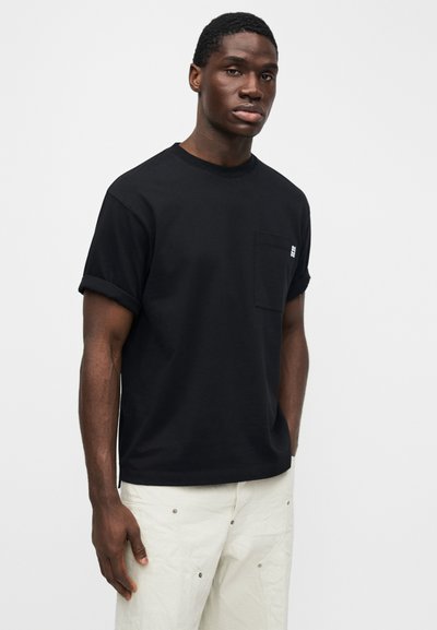 Camiseta negra de algodón con mangas remangadas y un bolsillo en el pecho izquierdo. Presenta un detalle de logo blanco minimalista en el área del bolsillo.