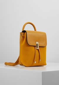 Żółty skórzany plecak z gładką górną klapą, złotymi okuciami i zamknięciem na sznurek. Posiada małą górną rączkę i regulowany pasek.