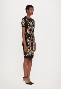 Rochie neagră cu mâneci scurte, prezentând un model floral colorat în nuanțe de roz și auriu. Este însoțită de tocuri negre cu vârf ascuțit și curea la gleznă.