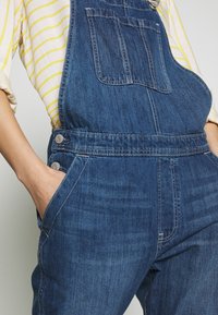 Denimbyxor i mörkblått med bröstficka, sidofickor, silverfärgad hårdvara och vita sömmar.