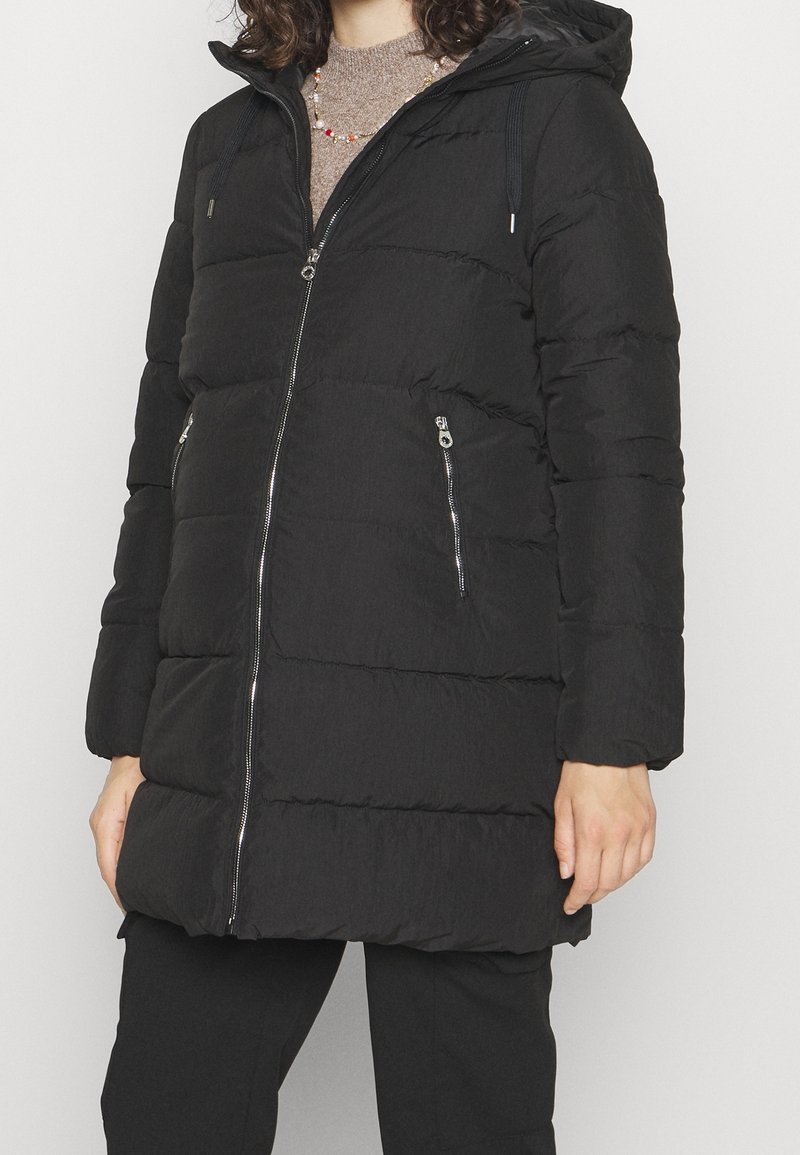 Personne portant un manteau noir matelassé à capuche avec une fermeture éclair frontale et des poches latérales zippées, superposé à un pull marron en tricot et un pantalon noir.