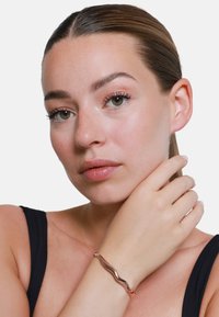 Bracciale in oro rosa con design ondulato, superficie liscia, indossato su un polso. Il modello ha un trucco neutro e accessori minimal.