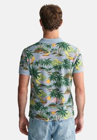 Poloko s krátkým rukávem světle modré barvy s tropickým potiskem zobrazujícím palm trees, pláže a plachetnice v zelených, žlutých a růžových odstínech.