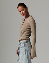 Μπεζ ριμπ μπλούζα με μακριά μανίκια, γιακά και διακοσμητικές βολάν στο μπροστινό μέρος. Συνδυασμένη με ανοιχτό μπλε τζιν παντελόνι με φαρδιά μπατζάκια.