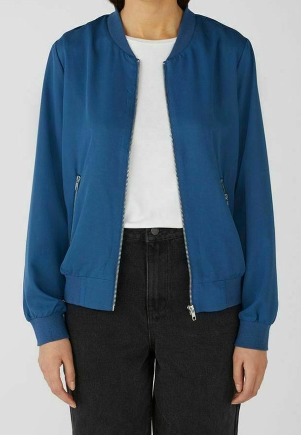 Femme portant une veste bomber bleue zippée sur une chemise blanche et un pantalon noir, debout devant un fond uni.