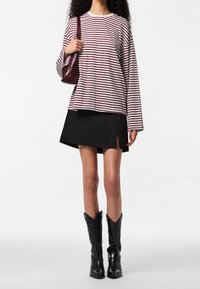 Femme vêtue d'un haut à manches longues rayé blanc et bordeaux, d'une mini-jupe noire avec une fente latérale, de bottes noires mi-mollet et tenant un sac à bandoulière bordeaux.