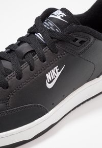 Zapatilla Nike de cuero negro con cordones negros, logo blanco, parte superior texturizada, cuello acolchado y suela de goma blanca con detalles en negro.