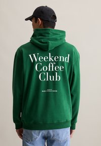 Man in een groene hoodie met witte tekst "Weekend Coffee Club" en een zwarte pet, staande voor een effen beige achtergrond.