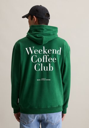 Mann in grünem Kapuzenpullover mit weißem "Weekend Coffee Club"-Text und schwarzer Mütze, stehen vor einem schlichten beigen Hintergrund.