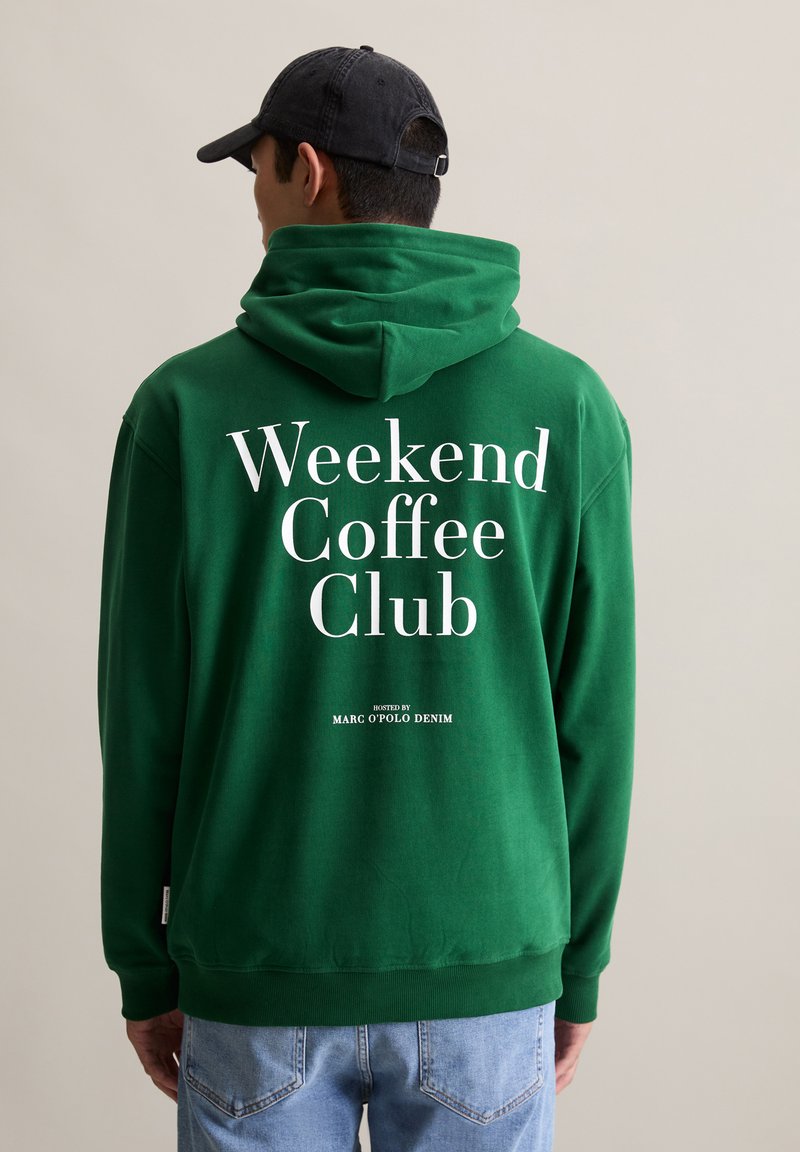 Man in een groene hoodie met witte tekst "Weekend Coffee Club" en een zwarte pet, staande voor een effen beige achtergrond.