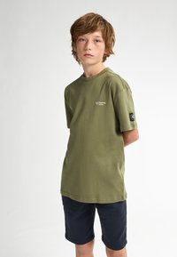 Camiseta de algodón verde oliva de manga corta, con un pequeño logo en el pecho y una etiqueta de acento negra en la manga.