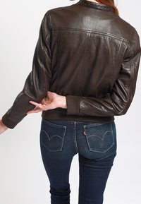 Brązowa skórzana kurtka o dopasowanym kroju, z prążkowanymi mankietami i przeszyciami z tyłu. Do niej ciemnoniebieskie dżinsy z kieszeniami w kształcie serc na plecach.