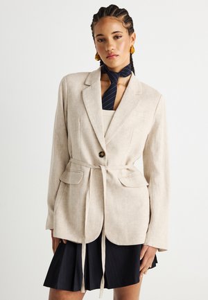 Γυναίκα με πλεξούδες στα μαλλιά, φορώντας μια μπεζ σακάκι με ζώνη, μαύρη πλισέ φούστα, ριγωτό μαύρο μαντήλι στο λαιμό και κίτρινα σκουλαρίκια, στέκεται μπροστά σε λευκό φόντο.