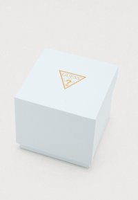 Weißer quadratischer Kasten mit goldenem Guess-Logo und dreieckigem Design oben, auf einer hellen Oberfläche platziert.