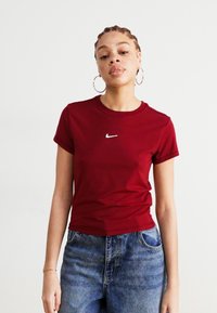 Burgundinės Nike marškinėliai trumpomis rankovėmis, su apvalia iškirpte ir baltu logotipu ant krūtinės, derinami su aukštu juosmeniu mėlynomis džinsomis.