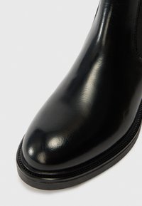 Musta kiillotettu nahkasaapas pyöreällä kärjellä ja paksulla pohjalla valkoista taustaa vasten.