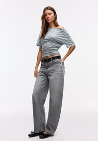 Donna con top a righe celesti e grigie scollo a barchetta, jeans grigi larghi a vita alta, cintura nera e ballerine nere, in posa su sfondo bianco.