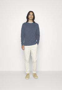 Blauer strukturierter Pullover mit gerippten Bündchen, cremefarbene Hose und hellbeige Schuhe. Das Model steht vor einem schlichten weißen Hintergrund.