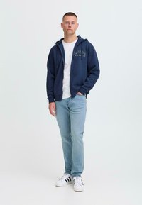 Mørkeblå zip-up hettegenser med frontlogo, brukt over en hvit t-skjorte, kombinert med lyseblå jeans og hvite sneakers med svarte striper.