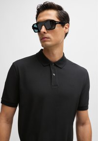 Black polo shirt with a classic collar, two buttons, and subtle branding. The individual wears square, dark sunglasses with blue lenses.
